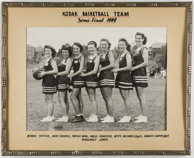 Schwarzes und weißes Foto einer Gruppe von Frauen, die lächeln und einen Basketball halten, mit der Aufschrift "Kodak Basketball Team Semi-Final 1949" oben und unten, vor einem Hintergrund aus Menschen, Bäumen, Gebäuden und dem Himmel.