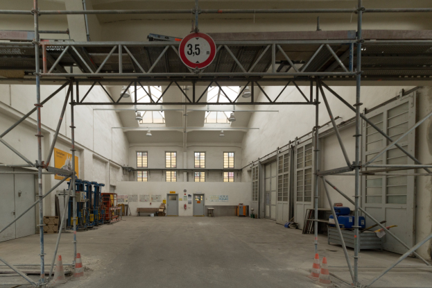 Lagerhalle mit Gerüsten, Verkehrskegeln, Metallstangen, einem Schild, Maschinen, einer Tür, Fenstern, Wänden, Deckenleuchten und einem Dach.