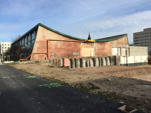 Großes grüner-dachtes Gebäude entlang einer Straße, umgeben von Bäumen, Pfählen und einem Zaun, mit bewölktem Himmel im Hintergrund; ehemaliges USK-Gebäude in Berlin, kürzlich renoviert.