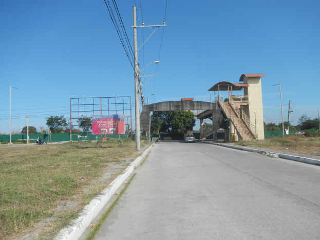 Straße mit Fahrzeugen, Strommasten, einem Schuppen mit Treppe und Geländer, einem "Zu verkaufen Grundstück / Land / Farm Cavite > Imus 1" Schild, Laternenmasten, Bäume und Himmel im Hintergrund.