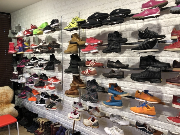 Ein Schuhgeschäft mit mehreren Ständern voller verschiedener Schuhe, einem Stuhl links mit einem Teddybär darauf und einer Wand im Hintergrund.