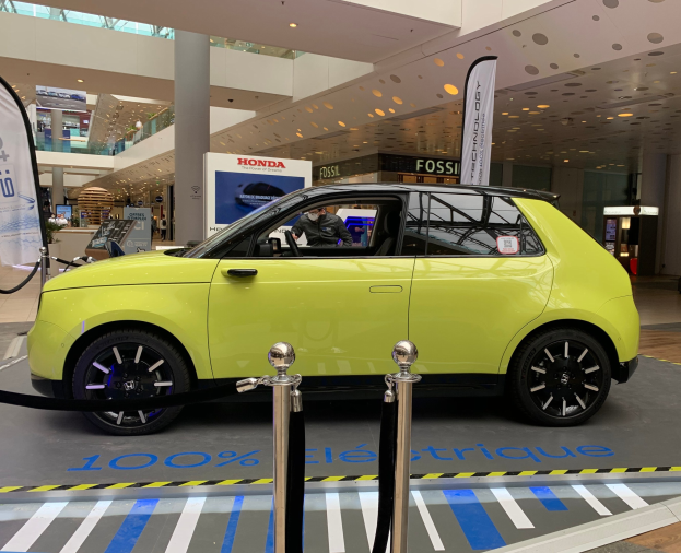 Gelber wasserstoffbetriebenes Elektroauto vor einem Gebäude mit Absperrpollern, Bannern, Menschen, Schildern, einer Topfpflanze, Deckenleuchten und Ventilatoren.