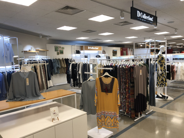 Innenansicht eines Kleiderladens mit Kleidern an Bügeln, Schaufensterpuppen und Tischen mit Artikeln, sowie Texttafeln an den Wänden und Deckenleuchten, identifiziert als der neue D Collection Store im Mall of America.