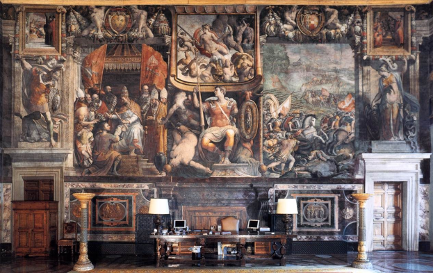 Großer Raum mit einem prominenten Gemälde an der Wand, ein Tisch mit Gegenständen, zwei Lampen, die den Tisch flankieren, zwei Türen an den Seiten und ein Teppich auf dem Boden, befindet sich im Palazzo della Signoria in Florenz, Italien.