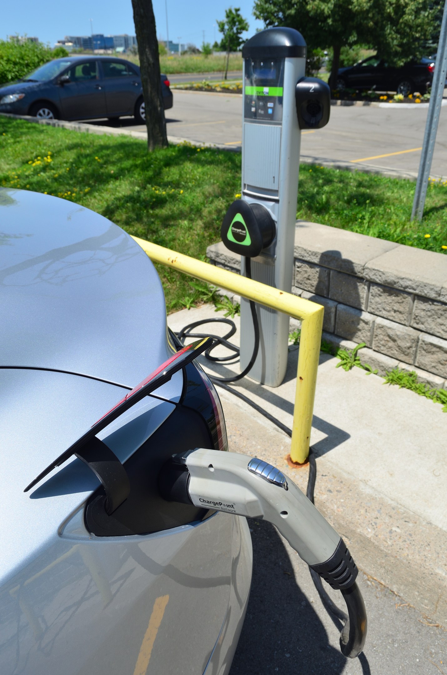 Elektrisches Auto an einer Straßenladestation mit Gras, Bäumen, Strommasten, Gebäuden und einem klaren blauen Himmel im Hintergrund.