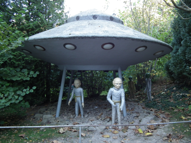 Gruppe von Menschen vor einem UFO mit Bäumen und einem klaren blauen Himmel im Hintergrund, Boden mit zerrissenen Blättern und Steinen bedeckt und ein Geländer im Vordergrund.