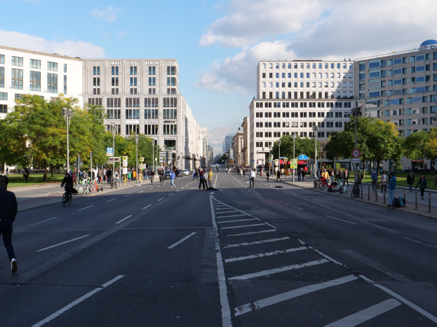 Eine belebte Straßenstraße in Berlin, Deutschland, mit Fußgängern und Radfahrern auf der Straße, hohe Gebäude mit Fenstern, Bäume, Laternenpfähle und Schilder, unter einem bewölkten Himmel.