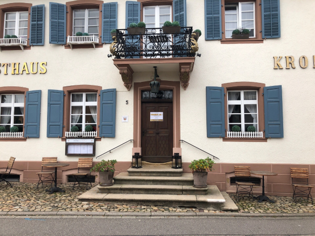 A building with blue shutters, windows, doors, railings, houseplants, tables, chairs, stairs, and a road in Sthaus, Germany.