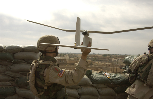 Zwei Soldaten in Helmen und Ausrüstung halten eine Drohne vor Sandsäcken, mit Gebäuden, Bäumen und einem bewölktem Himmel im Hintergrund.