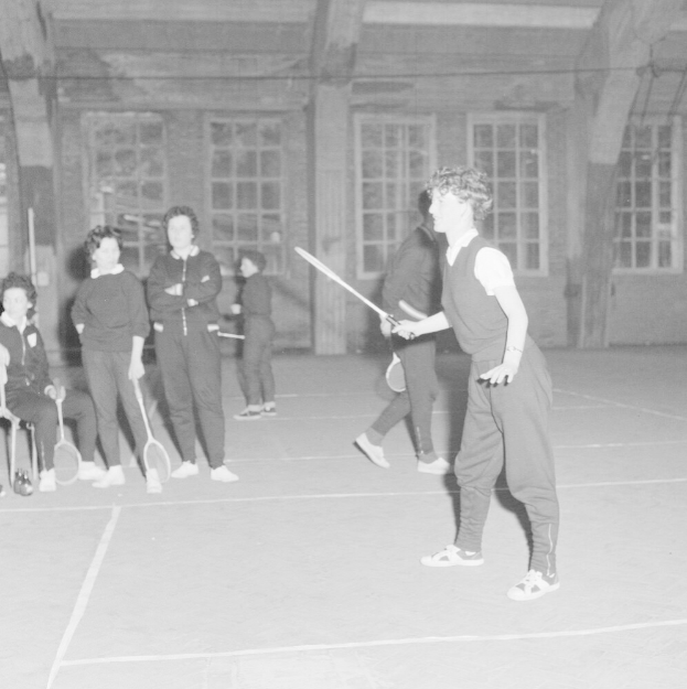 Schwarz-weißes Bild von Menschen, die Badminton auf einem Platz spielen, einige halten Schläger, mit Zuschauern auf Stühlen links und einer Wand mit Fenstern im Hintergrund.