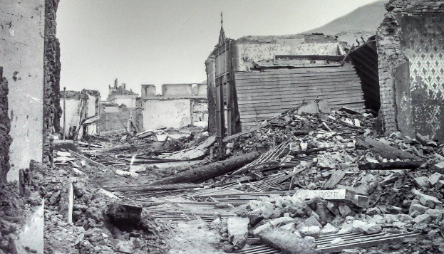 Ein Schwarz-Weiß-Foto eines zerstörten Gebäudes in der Mitte einer Straße, umgeben von eingestürzten Wänden, Holzstöcken, Steinen und anderem Schutt, mit einem Hügel und dem Himmel im Hintergrund, der auf die Zerstörung hinweist, die durch das Erdbeben verursacht wurde, das 1945 in Mexiko-Stadt stattfand.
