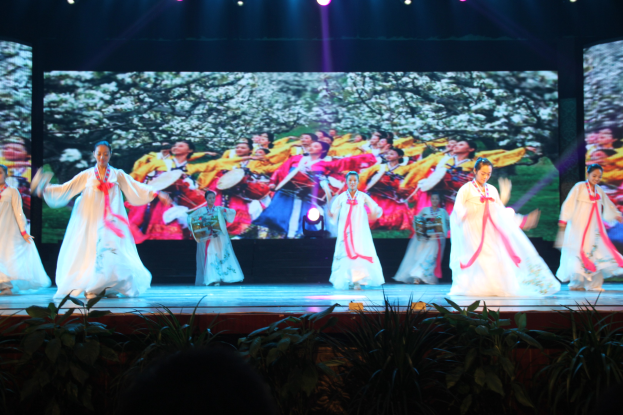 A group of people performing a traditional Korean dance on a stage with lighting and plants, with a large screen in the background.