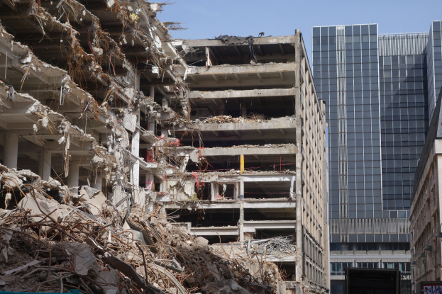 Large building demolished in an urban area with surrounding structures, vehicles, street elements, trees, and clear sky.
