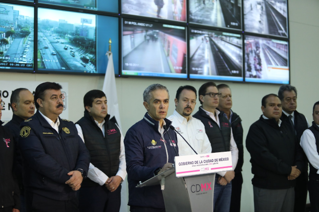 Gruppe von M├Ąnnern, die vor einem Podium mit einem Mikrofon und einer Tafel mit der Aufschrift 'Mexikanische Zoll- und Grenzschutzbeh├Ârde' stehen, mit Fernsehern und einer Flagge an der Wand dahinter.