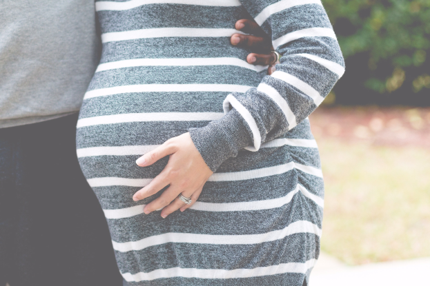 Schwangere Paare in Schwangerschaftskleidung, das zusammen mit den Händen auf dem Bauch steht, umgeben von Bäumen.