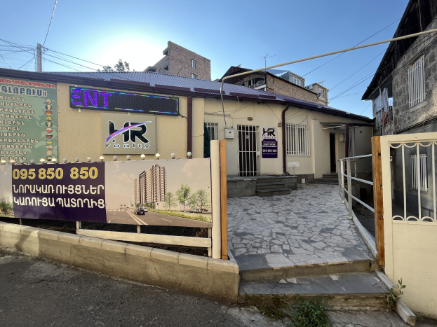 Gebäude mit einem Schild, das "hr Immobilienagentur-Büro zur Miete in Phnom Penh" trägt, vorne, umgeben von einem Metallzaun und einem Tor, mit umgebenen Pflanzen, einem Weg, Polen, Drähten, Bäumen und einem bewölkten Himmel.