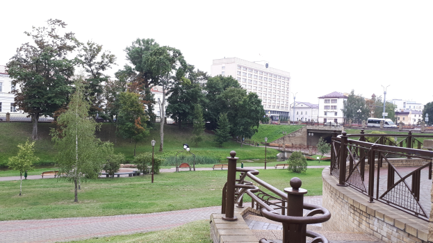 Eine Parkszene mit einer Brücke über einen Fluss, umgeben von Gebäuden, Bäumen, Straßenlaternen, Straßenlaternen, Kraftfahrzeugen, Geländern, Bänken, Pflanzen, Gras und einem klaren blauen Himmel.