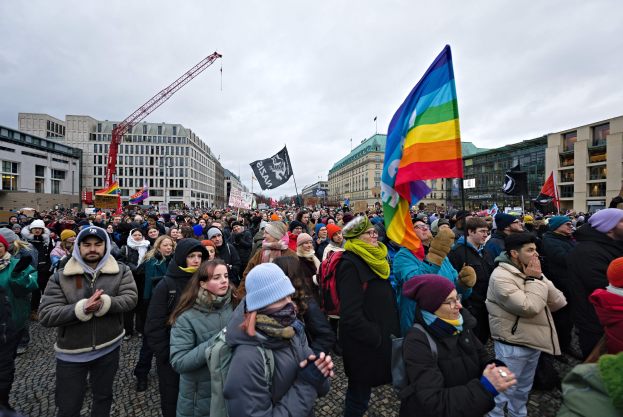 Eine große Gruppe von Menschen steht vor einem Gebäude, hält Fahnen und Schilder mit Text, darunter eines mit der Aufschrift 'Lgbtq+ Rechte Demonstrationen in Berlin', während einige Mützen tragen und Taschen tragen; Gebäude, ein Kran und ein bewölkter Himmel sind im Hintergrund zu sehen.
