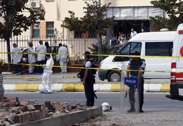 Gruppe von Polizeibeamten in der Nähe eines beschädigten Gebäudes mit Trümmern, einschließlich Ziegelsteinen und einem Helm auf dem Boden, umgeben von Fahrzeugen, einem Zaun und Vegetation.