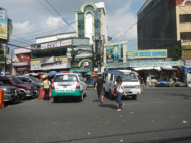 Eine belebte Straße in Manila, Philippinen, mit Fußgängern, Fahrzeugen, Schirmen, Gebäuden, Schildern, Strommasten, Bäumen und einem bewölkten Himmel, mit einem geparkten Polizeiwagen im Vordergrund.