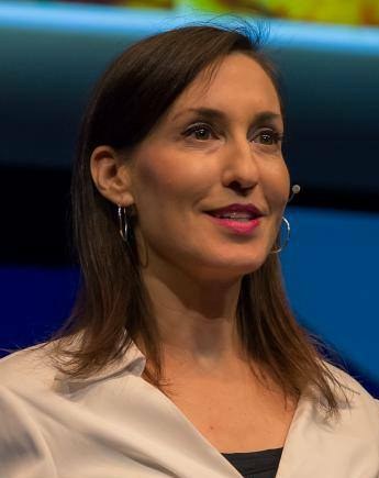 Frau in weißer Jacke und schwarzem Oberteil, die vor einem blauen und gelben Hintergrund sitzt und Ohrringe und ein Mikrofon trägt.