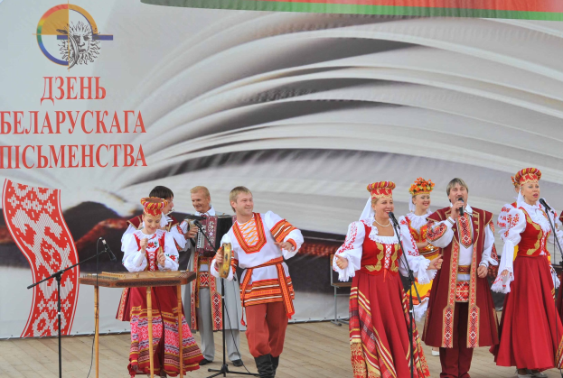 Eine Gruppe von Menschen in rot-weißer Kleidung tritt auf der Bühne mit Mikrofonen und Instrumenten auf, mit einem "Ukrainischer Folklore-Festival"-Schild im Hintergrund.
