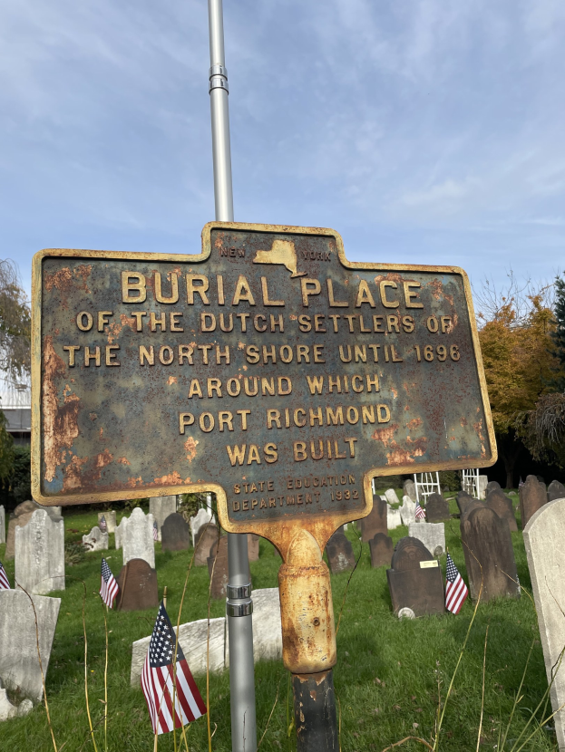 Grabanlage niederländischer Siedler an der Nordküste mit Texttafel, Fahnenmast, Gräbern, Bäumen, einem Gebäude und einem bewölkten Himmel.