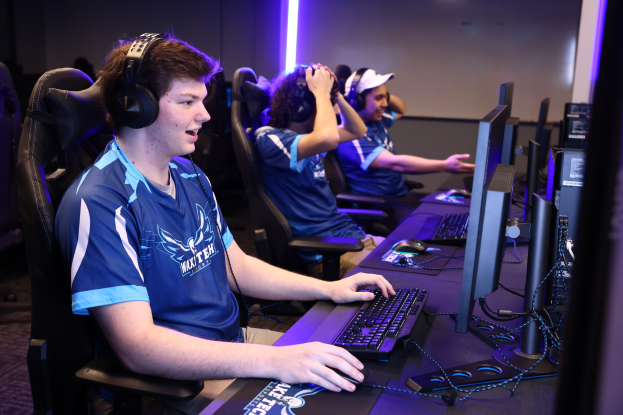 Gruppe junger Männer mit Headsets, die ein Videospiel auf Computern mit Monitoren, Tastaturen und Mäusen auf einem Schreibtisch spielen, mit beleuchteter Wand im Hintergrund.