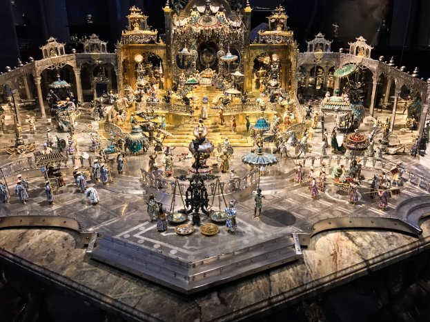 Eine große Gruppe von Menschen in bunten Kostümen auf einer hell erleuchteten Bühne umgeben von Spielzeugen, Skulpturen und einem gerahmten Fotohintergrund aus *Der Nussknacker und die vier Reiche*.