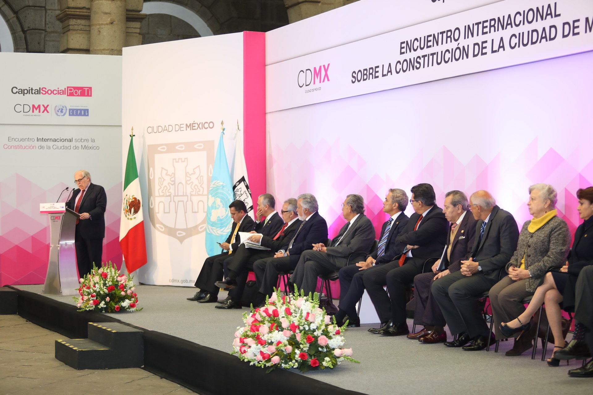 Gruppe von Menschen, die auf Stühlen sitzen und einem Podium mit Mikrofon zugewandt sind, umgeben von Blumengestecken, Flaggen und Texttafeln, vor einem Hintergrund aus Säulen und einer Wand.