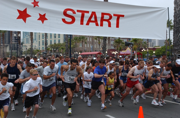 Gruppe von Menschen, die bei einem Marathon laufen, mit einem Verkehrskegel im Vordergrund und einem Banner im Hintergrund, umgeben von Bäumen, Laternen, Gebäuden und einem klaren blauen Himmel.