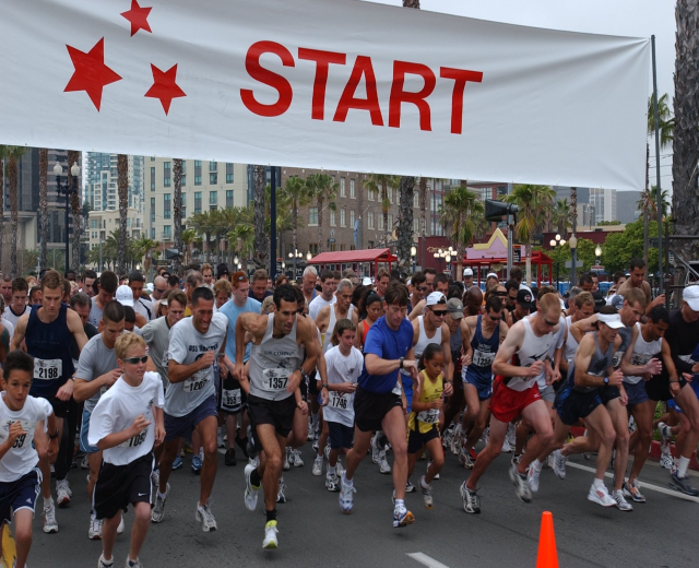 Gruppe von Menschen, die bei einem Marathon laufen, mit einem Verkehrskegel im Vordergrund und einem Banner im Hintergrund, umgeben von Bäumen, Laternen, Gebäuden und einem klaren blauen Himmel.