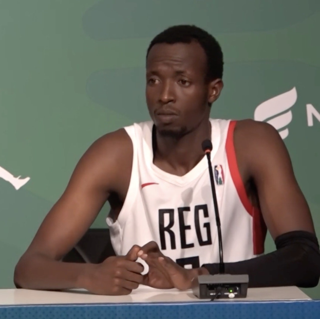 Ein Mann in einem weißen und roten Kleid sitzt an einem Tisch mit einem Mikrofon, vor einer grünen Tafel mit der Aufschrift "NBA All-Star Game Press Conference", mit einem Gegenstand auf dem Tisch dahinter.