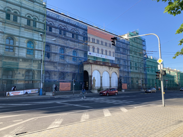 Großes Gebäude mit Gerüst während der Renovierung, umgeben von Fahrzeugen, Fußgängern, Ampeln, Straßenschildern, Pfosten, Bäumen und einem klaren blauen Himmel.