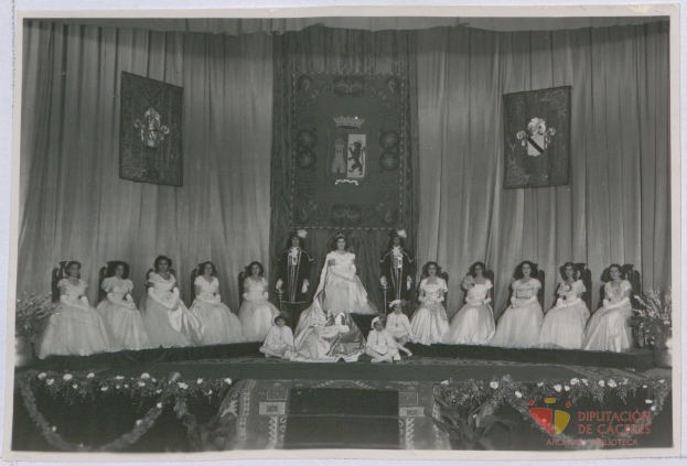 Gruppe von Frauen in weißen Kleidern auf einer Bühne sitzend mit Vorhängen und gerahmten Gegenständen an der Wand dahinter und Text am unteren Bildrand.