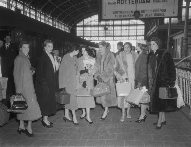 Schwarz-weißes Bild von Frauen, die an einem Bahnhof stehen und Taschen halten, neben einem Zug auf der linken Seite und Geländern mit einer Tafel auf der rechten Seite.
