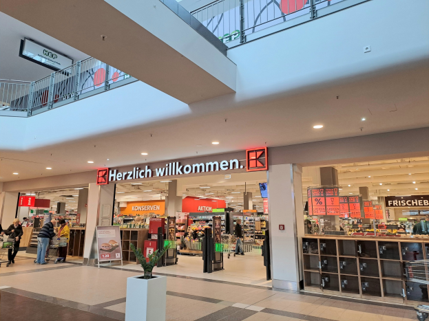 Interior of a shopping mall with people, stores, illuminated signs including "Herzlich willkommen" (German for "Welcome"), railings, overhead lights, and a potted plant at the bottom.