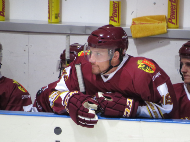 Gruppe von Eishockeyspielern, die auf einer Bank sitzen und Helme und Handschuhe tragen, mit Dosen und Gegenständen im Hintergrund auf einem Regal.