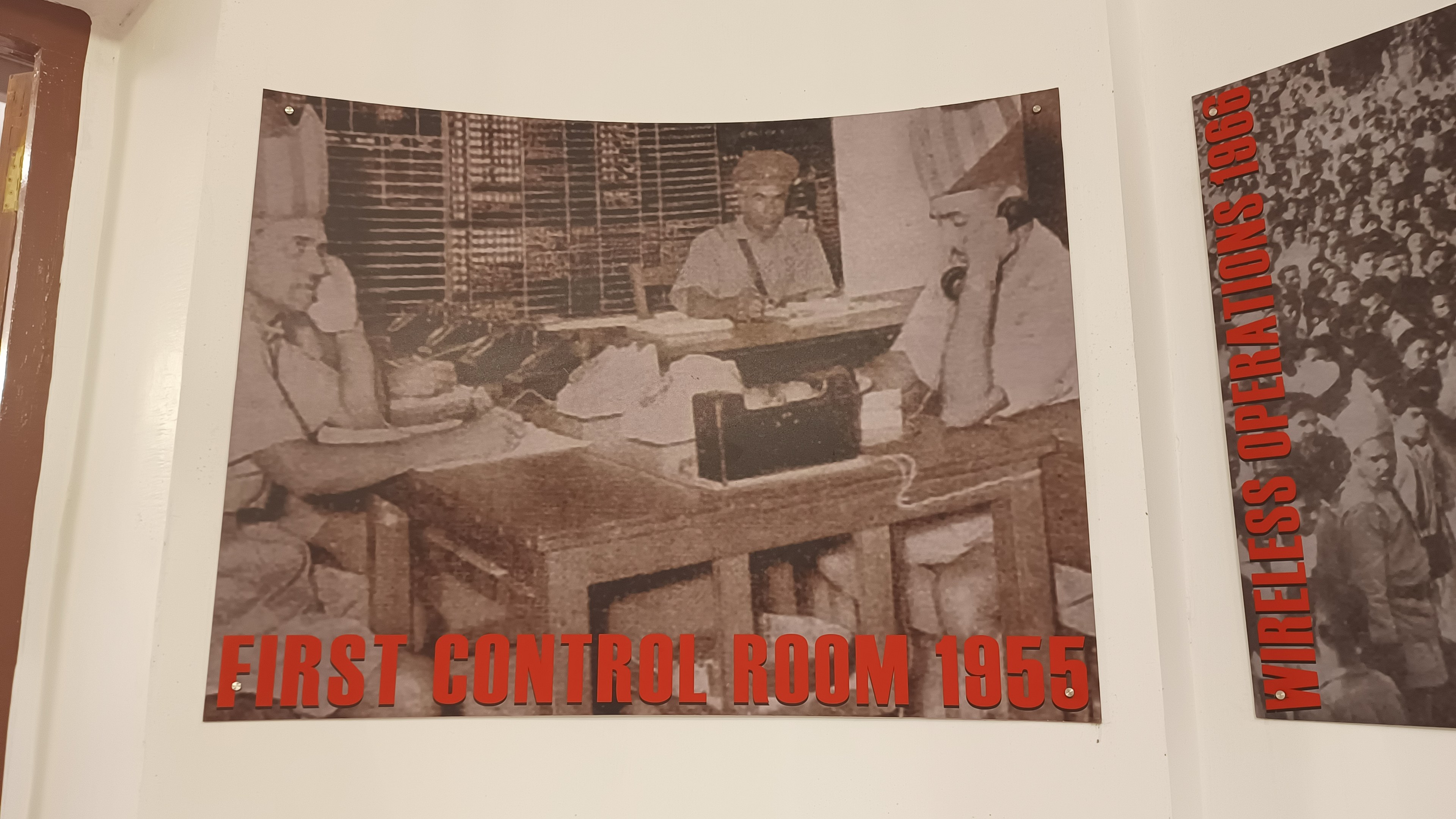Eine Wand mit zwei Postern, beschriftet mit "First Control Room 1955" und "Wireless Operations", ein zentraler Tisch mit Gegenständen und eine Tür auf der linken Seite.