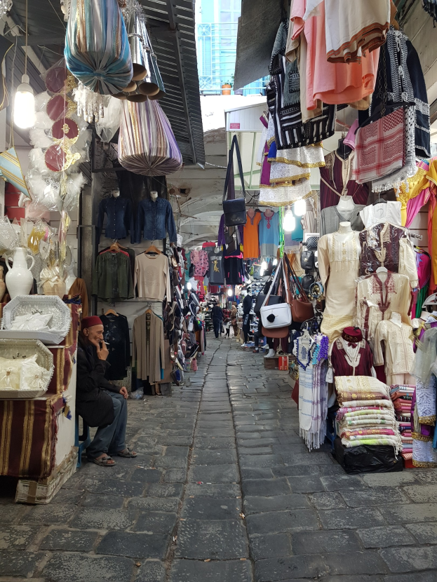 Ein belebter Straßenmarkt in Jerusalems alter Stadt mit Ständen, die Kleidung, Taschen, Lampen und andere Gegenstände verkaufen, umgeben von Menschen, die durch die Gassen schlendern und ein Gebäude im Hintergrund.