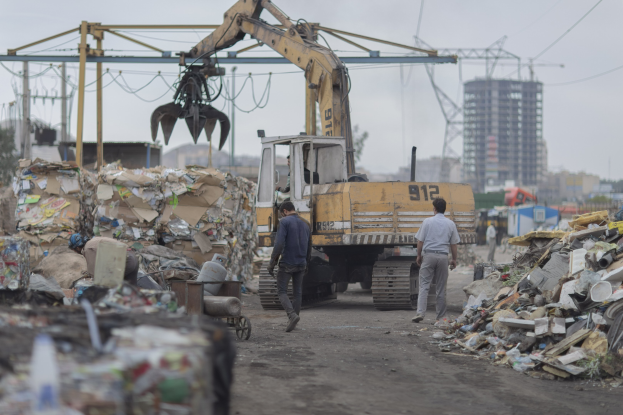 Eine Gruppe von Männern, die auf einem Schotterweg neben einem Mülllhaufen gehen, mit einem Bagger und Gebäuden im Hintergrund.