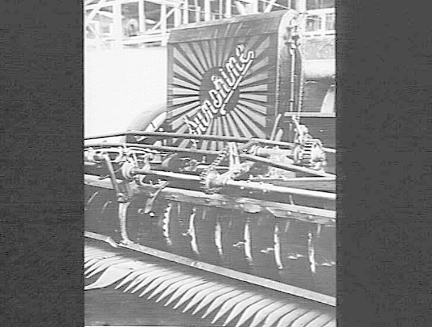 Schwarzes und weißes Foto einer Mähmaschine in einer Fabrikeinrichtung mit "Mähmaschine"-Text darauf und einem Gebäude im Hintergrund.