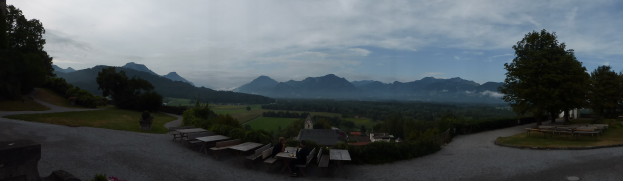 Panoramablick auf ein Gebirge von einem Hügel aus mit Menschen, Bänken, Tischen, blühenden Pflanzen, Gras, einem Zaun, Häusern, Bäumen und einer bewölkten Himmel.