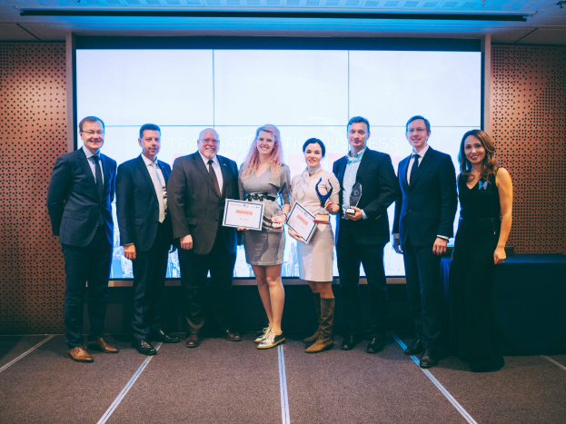 Gruppe von strahlenden Menschen mit Urkunden vor einem Bildschirm, mit einem schwarzen Tischtuch und einem wandmontierten Objekt im Hintergrund, Gewinner der International Business Awards 2018.