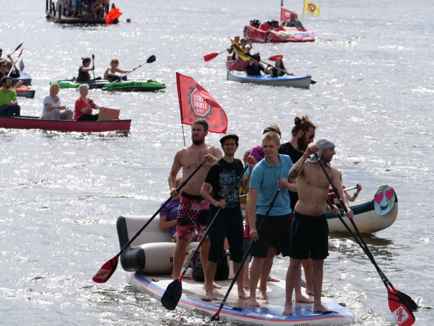Eine Gruppe von Menschen, die auf einem Paddleboard im Wasser stehen, einige halten Paddel, mit Booten und Fahnen im Hintergrund während des Stand Up Paddleboard Festivals 2016.
