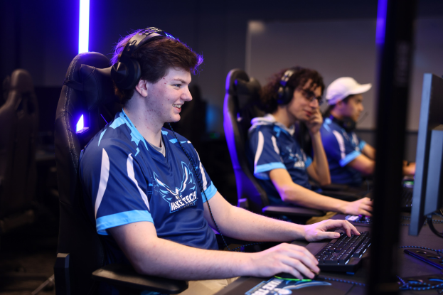 Eine Gruppe junger Männer in Headsets lächelt, während sie an einem Computersystem sitzen, wahrscheinlich ein E-Sport-Spiel, mit Monitoren, Tastaturen und anderen Gegenständen auf dem Schreibtisch und Lichtern im Hintergrund.