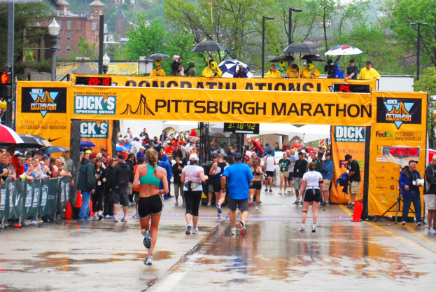 Eine Gruppe von Läufern nimmt an einem regnerischen Tag am Pittsburgh-Marathon teil, einige halten Schirme, mit Absperrungen, Verkehrskegeln, Bannern, Laternenpfählen, Bäumen, Gebäuden und einem klaren Himmel.