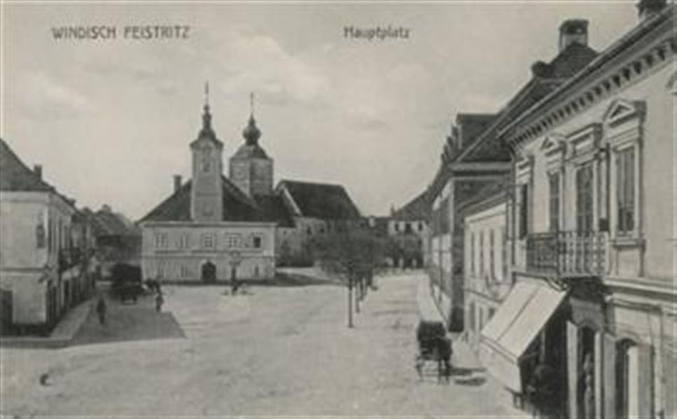 Black and white photo of a city street in Windisch Feststritz, Germany, featuring buildings, trees, people, carts, and a sky in the background.