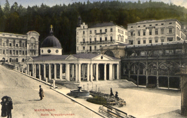 Schwarz-weiß-Foto eines Stadtplatzes mit einem zentralen Brunnen, umgeben von Gebäuden, Menschen, Bäumen und einem bewölkten Himmel, mit Text am unteren Rand.
