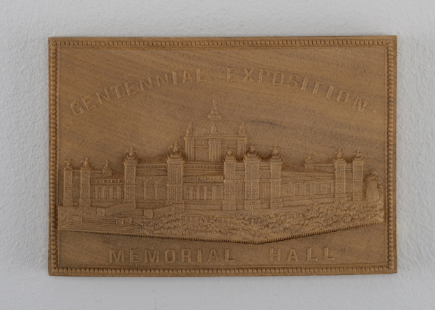 Holzschild an einer Gebäudewand mit der Aufschrift "Centennial Exposition Memorial Hall" und einem eingravierten Bild des Gebäudes.